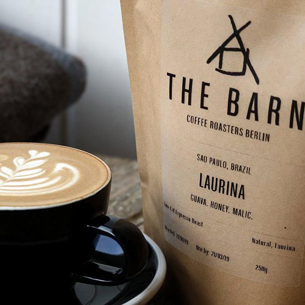 A bag of Laurina, Brazil with a cup of coffee with a latte-art design, both on a wooden table